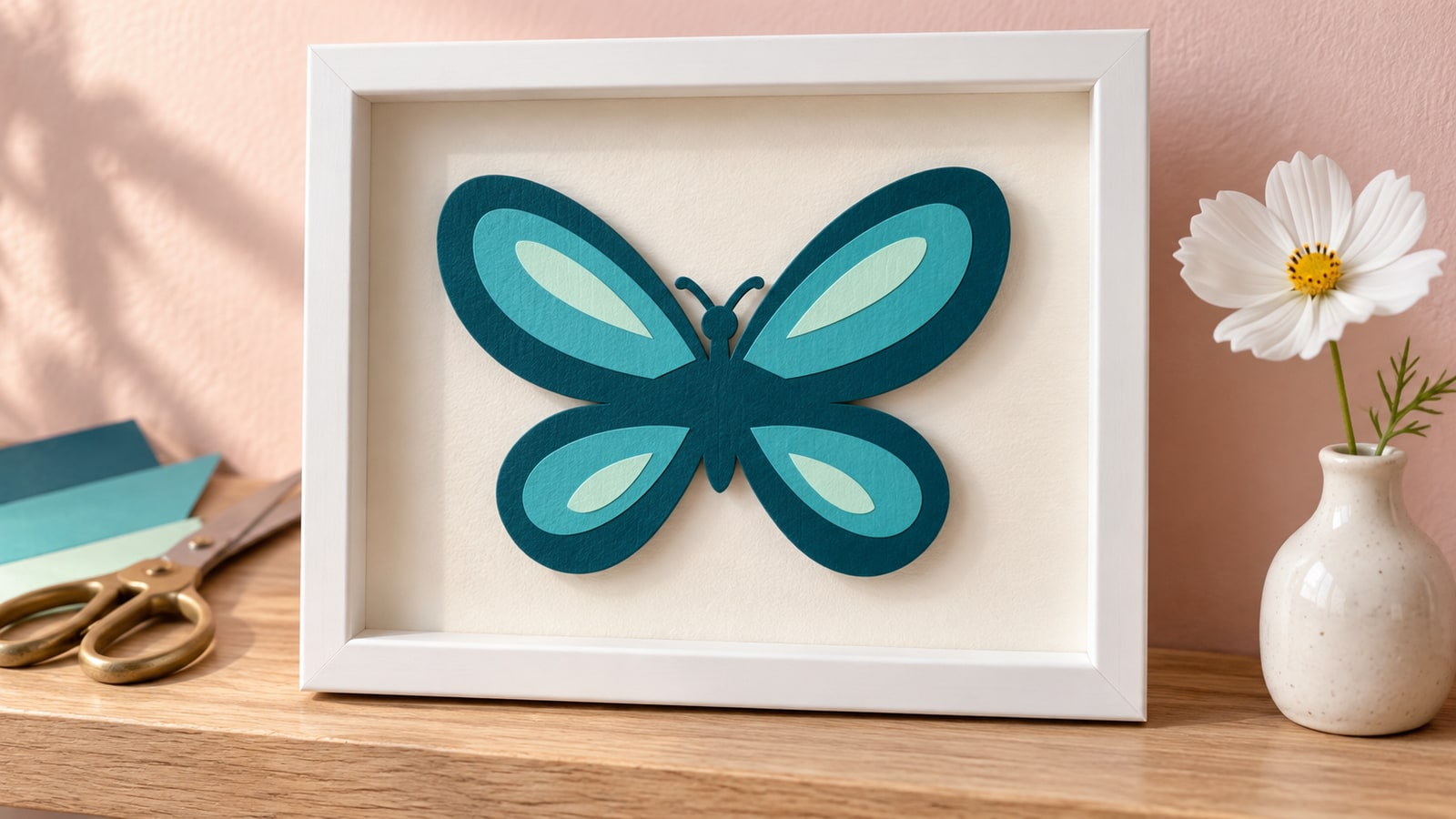Teal, aqua and mint paper butterfly in a white shadow box on an oak shelf beside a white flower